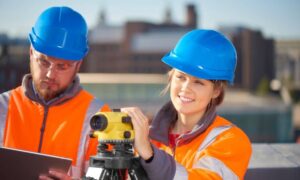Surveyors conducting an ALTA land title survey, wearing blue helmets and orange safety vests while using surveying equipment on-site
